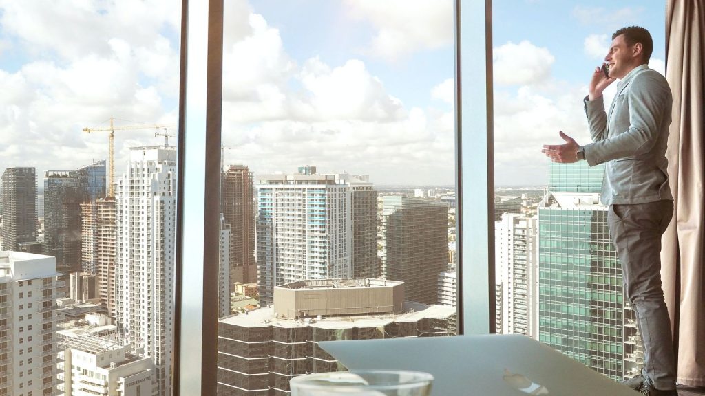 Affaires qui ralentissent ? Peur de la prospection ? Manque de compétences ?  Doit-on prospecter lorsqu’on est dirigeant d’une petite entreprise ? Homme d'affaire, téléphone à la main, dans son bureau, devant une baie vitrée qui donne sur des bulidings