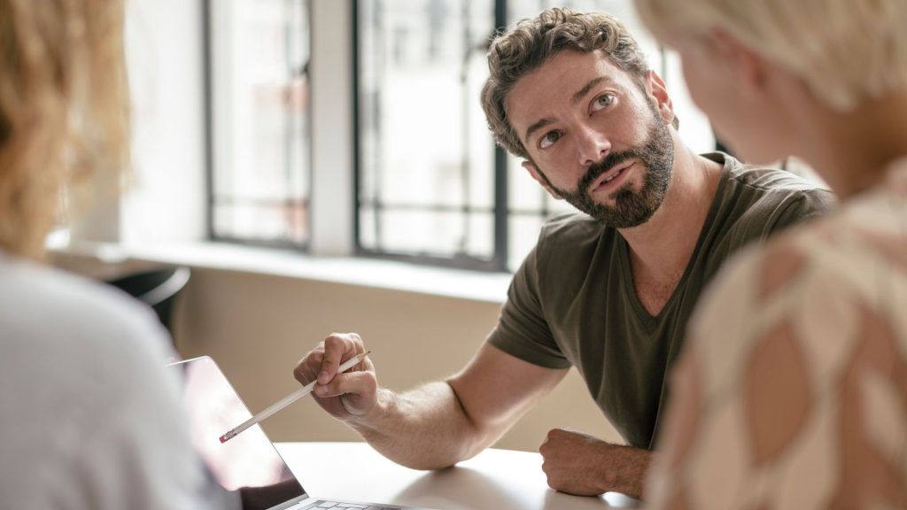 Homme mature avec barbe pointant sur ordinateur portable avec un crayon et à l’écoute de collègue