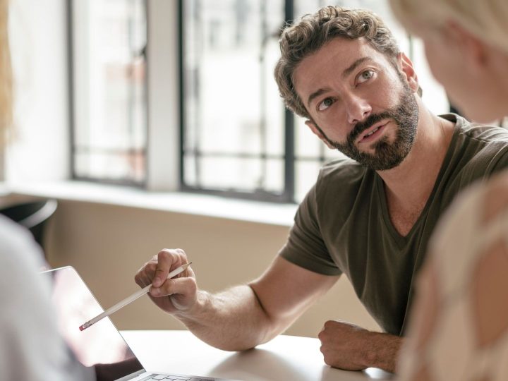 Homme mature avec barbe pointant sur ordinateur portable avec un crayon et à l’écoute de collègue