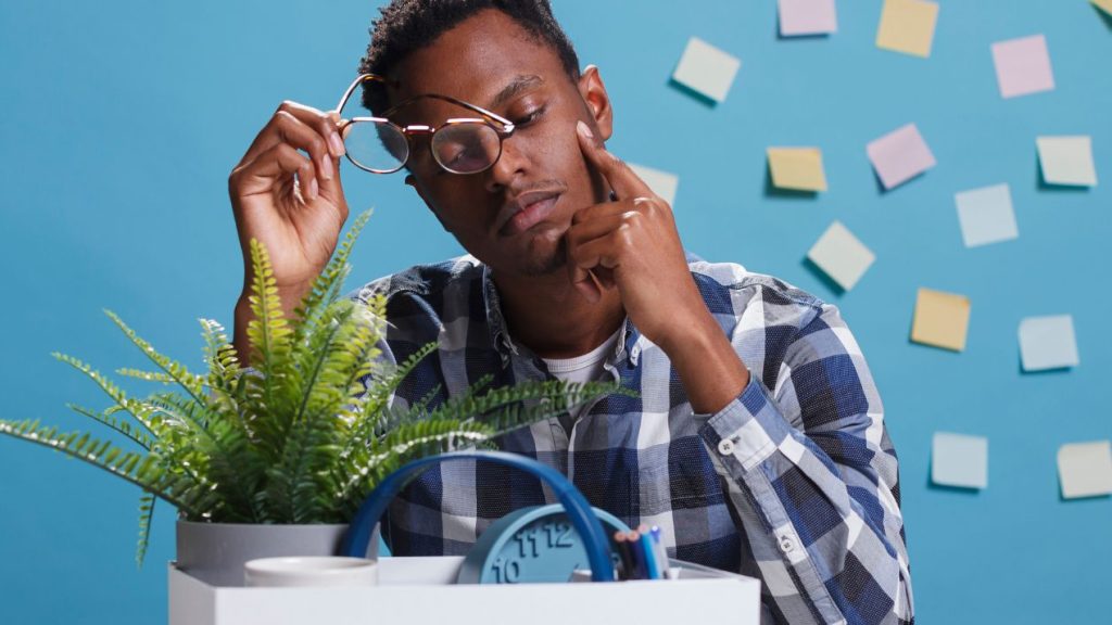 Le rapport EPOS « Workplace of the Future » explore les tendances mondiales qui façonneront le travail hybride en 2023. jeune adulte epuisé et pensif avec un carton contenant ses affaires professionelles posées sur le bureau et des post-it sur le mur